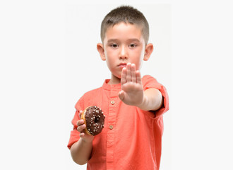 Sticker - Dark haired little child eating doughnut with open hand doing stop sign with serious and confident expression, defense gesture