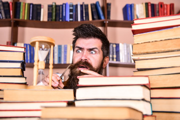 Leinwandbilder - Man, scientist looks at hourglass. Man on serious and shocked face watching time is going over, bookshelves on background. Time flow concept. Teacher or student with beard studying in library.