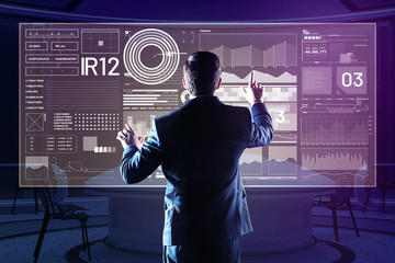Comparing diagrams. Professional web developer standing in front of his futuristic computer and carefully touching the transparent screen while working
