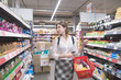 © bodnarphoto - Portrait of a beautiful girl walking in a supermarket with a shopping cart in her arms and looking for products. Stylish young woman shopping in a supermarket.