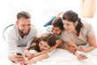 © Drobot Dean - Portrait of joyful family with two children laughing, and taking selfie on smartphone while lying together on bed in apartment