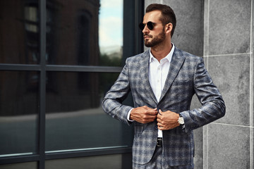 Portrait of sexy handsome fashion businessman model dressed in elegant checkered suit posing near gray wall on street background. Metrosexual