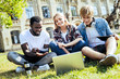 © Viacheslav Yakobchuk - Dream team. Three joyful students relaxing on grass and smiling while focusing their attention on a screen of a laptop.