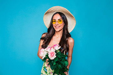 Wall Mural - Happy and smiling young woman portrait with long curly hair in summer dress with bouquet of flowers in hands posing over blue background. Holidays and gifts concept