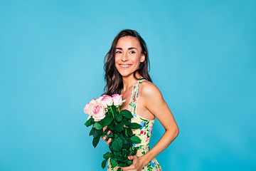 Wall Mural - Happy and smiling young woman portrait with long curly hair in summer dress with bouquet of flowers in hands posing over blue background. Holidays and gifts concept