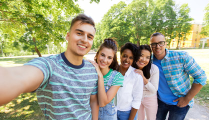 Wall Mural - people, friendship and international concept - happy smiling young woman and group of happy friends taking selfie outdoors