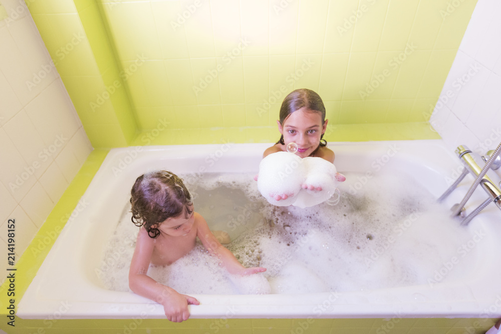 Happy laughing sister kids taking a bath playing with foam bubbles. Little children in a bathtub. Family lifestyle indoors