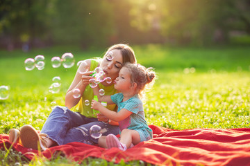 family in park -female child blows soup foam and make bubbles with her mother in nature.