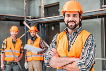Handsome Smiling Builder Standing At Construction Site With Crossed Arms While His Colleagues Standing Blurred On Background