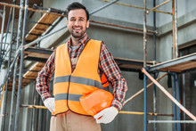 Happy Builder In Reflective Vest Holding Helmet While Standing In Front Of Building House And Looking At Camera