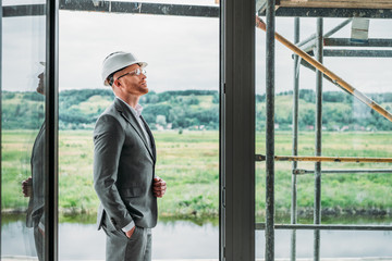 Wall Mural - side view of handsome architect in suit and hard hat standing on terrace at construction site