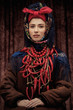 Portrait of young pretty woman sitting in ethnic winter costume with hands on her lap, wearing warm coat, shawl, knotted headband, beads and paprika necklace, looking at camera over brown background