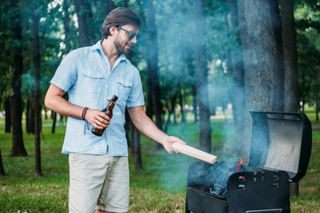 smiling man in sunglasses with beer setting fire on grill during barbecue in park