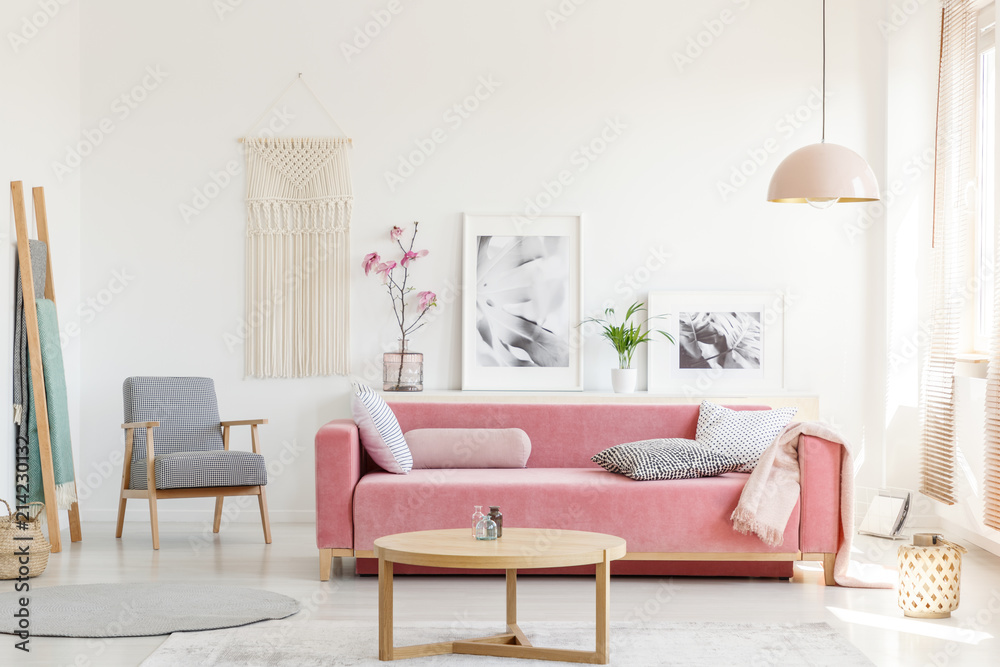 Patterned armchair next to pink sofa in apartment interior with posters and wooden table. Real photo - obrazy, fototapety, plakaty 