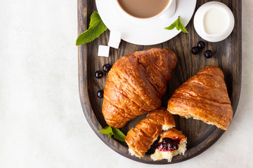 Breakfast concept: two fresh french croissant with a cup of coffee and jam on a dark wooden tray.