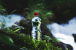 © yanik88 - Transparent plastic A bottle of clean water with a red lid stands in the grass and moss on the background of a rugged mountain river. The concept of pure natural drinking water