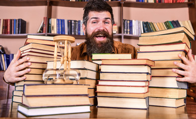 Leinwandbilder - Man on happy face between piles of books, while studying in library, bookshelves on background. Librarian concept. Teacher or student with beard sits at table with books, defocused.