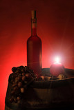 Fototapeta  - Butelka czerwonego wina w świetle świecy na beczce z winogronem.