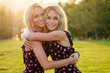 two sisters twins beautiful curly blonde happy young toothy smile women in stylish dress hugging in the summer park sunset rays field background