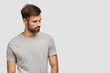 © WHstudio Leushin N - Indoor shot of concentrated handsome unshaven young male foused aside, has muscular body, dressed in casual grey t shirt, stands against white background with blank space for your advertisement