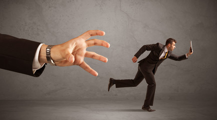 Wall Mural - Young miniature businessman running from a big hand with grey background