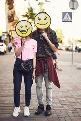 Wall Mural - Full length portrait of beaming lady and happy male with cartoon faces embracing while going along city during sunny day