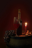 Fototapeta  - Butelka czerwonego wina w świetle świecy na beczce z winogronem.