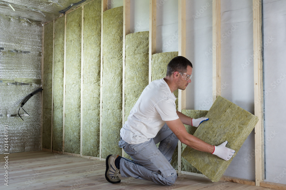 Worker in protective goggles and respirator insulating rock wool insulation in wooden frame for future house walls for cold barrier. Comfortable warm home, economy, construction and renovation concept - obrazy, fototapety, plakaty 