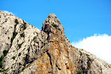 A Sheer Wall Of Rock With Scanty Green Vegetation And A Ledge In The Middle Against A Blue Sky With A Floating Cloud.