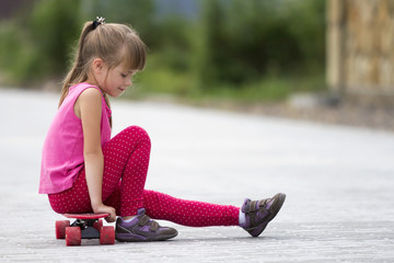 Pretty young long-haired blond child girl in casual pink clothing sitting on skateboard on paved suburb street on blurred sunny summer green background. Children activities, games and fun concept.