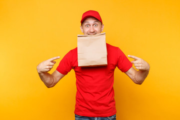 Man in red cap, t-shirt giving fast food order isolated on yellow background. Male employee courier hold in mouth paper packet with food. Products delivery from shop or restaurant to home. Copy space.