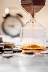 Sand running through the shape of hourglass on table with banknotes and coins of international currency. Time investment and retirement saving. Urgency countdown timer for business deadline concept