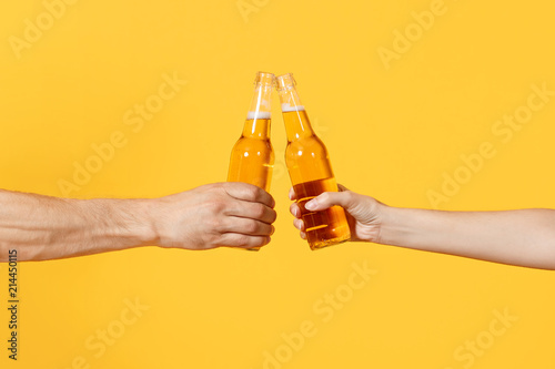 Fotografía Close up cropped of woman and man two hands horizontal holding lager beer glass bottles and clinking isolated on yellow background