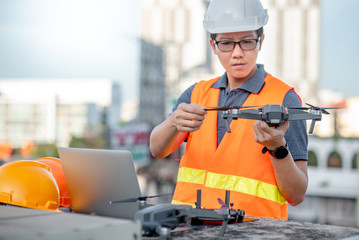 Young Asian engineer man working with drone laptop and smartphone at construction site. Using unmanned aerial vehicle (UAV) for land and building site survey in civil engineering project.