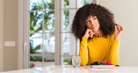 Poster - African american woman eating raspberries and blueberries at home serious face thinking about question, very confused idea