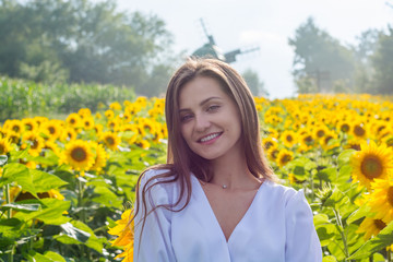 Beautiful smiling young woman in a white shirt stands in the field among sunflowers at a rural landscape background