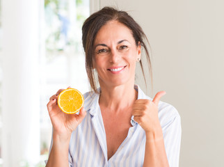 Middle aged woman holding orange fruit happy with big smile doing ok sign, thumb up with fingers, excellent sign