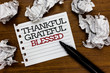 © Artur - Text sign showing Thankful Grateful Blessed. Conceptual photo Appreciation gratitude good mood attitude Paper lumps laid randomly around white notepad touch black pen on woody floor.