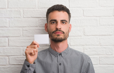 Young adult man over brick wall holding blank card with a confident expression on smart face thinking serious