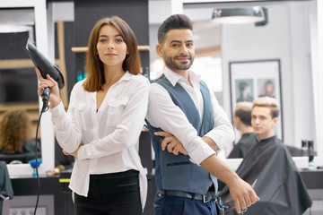 Two hairstylers posing standing in modern beaty salon, young client sitting behind. Hairdressers wearing classic white shirts, man having grey waistcoat. Woman keeping hair dryer, looking at camera.