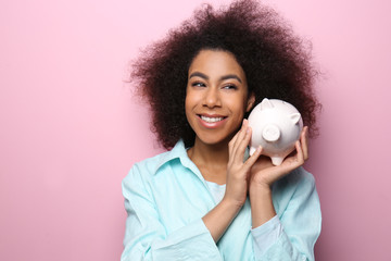 Canvas Print - Young African-American woman with piggy bank on color background. Savings concept