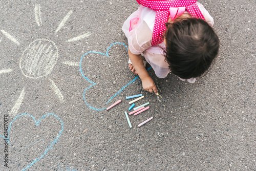 Fotografía Top view image of happy little girl wears pink dress and backpack drawing with colorful chalks on the sidewalk
