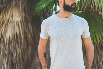 Hipster handsome male model with beard wearing gray blank t-shirt and a black snapback cap with space for your logo or design in casual urban style.Green palm and cactus garden on the background