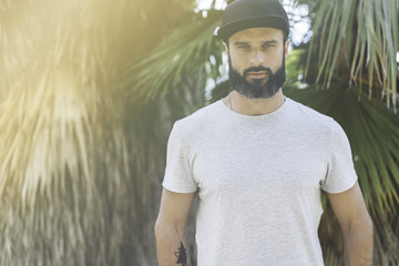 Wall Mural - Hipster handsome male model with beard wearing gray blank t-shirt and a black snapback cap with space for your logo or design in casual urban style.Green palm and cactus garden on the background