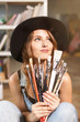 © proimagecontent - Portrait of pretty romantic long-hair female artist in cute black hat holding many brushes in contemporary studio