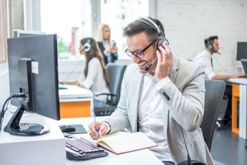 Wall Mural - Businessman taking notes while talking with customer using headphones and microphone in call center