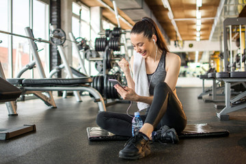 Cheerful young woman reading good news on mobile phone and celebrating success during workout break in gym