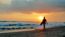 Slow Motion Video Of Young Male Surfer Walking On Sandy Beach With Surfboard At Summer Sunset Time. Bright Orange Sun Shines Over Horizon Of Bali Island In Indonesia