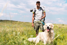 Male Traveler Walking With Golden Retriever Dog On Summer Meadow With Cloudy Sky