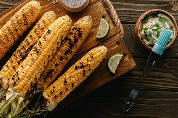 top view of cutting board with grilled corn, lime slices near cooking brush and butter with parsley on wooden table